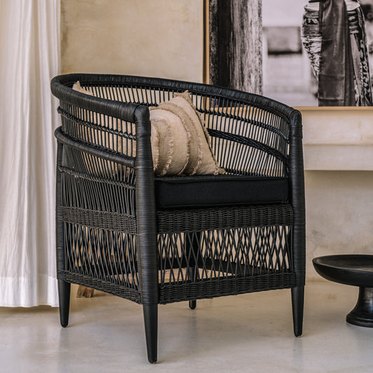 A black rattan chair with a handwoven design, featuring a cushion on the seat, positioned in a room with a light curtain background.