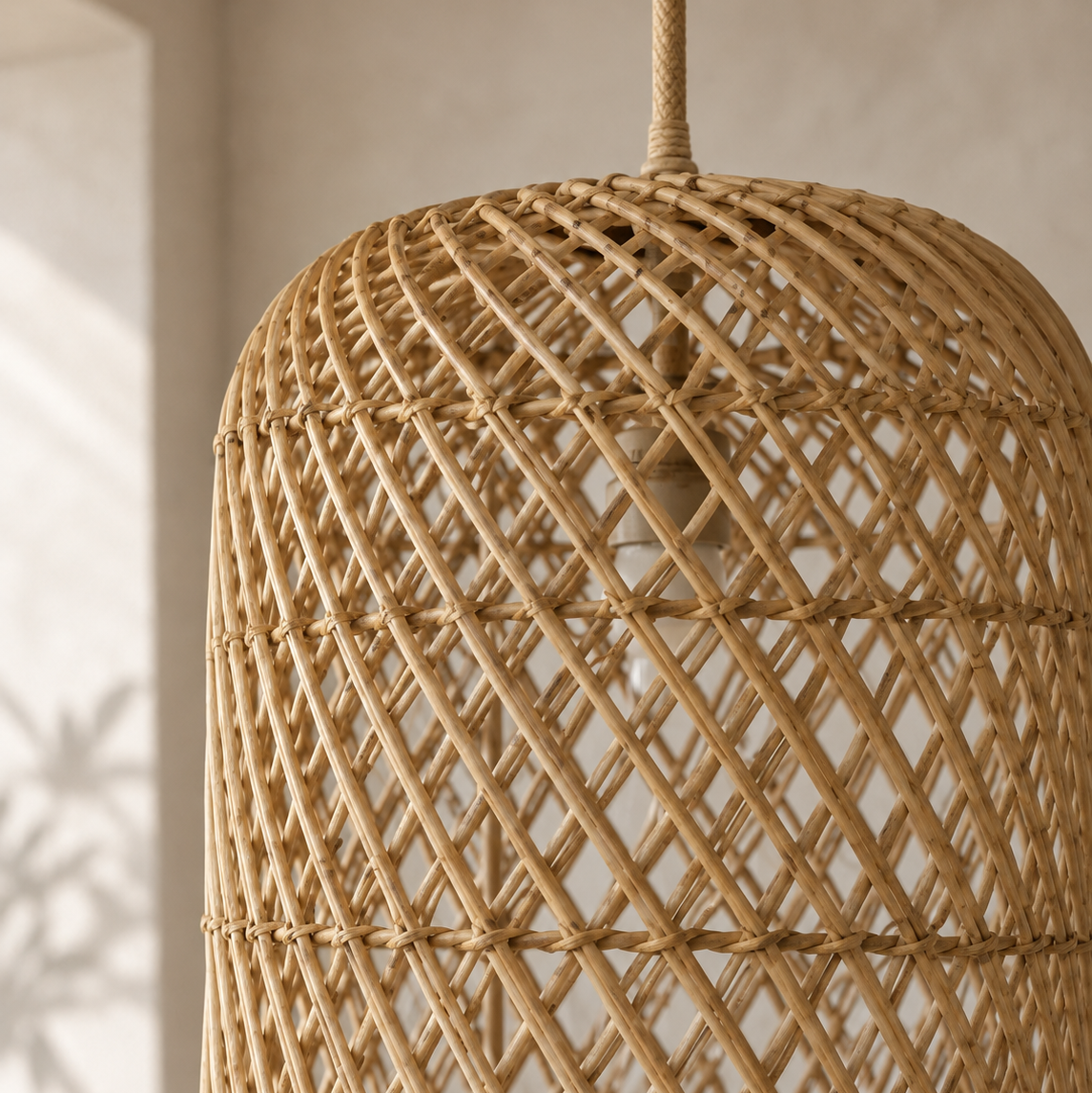 Close-up of a woven natural rattan pendant light in a warm tan color, hanging against a softly lit neutral wall.