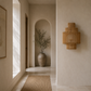 A modern tiered wall sconce made of natural woven rattan, mounted on a textured beige plaster wall in a minimalist hallway with a woven rug and a potted plant in the background.