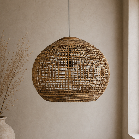 A spherical woven rattan pendant light hangs from a black cord against a warm beige plaster wall, with a large ceramic vase and dried stems on the left and a window light on the right.