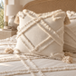A cream-colored textured throw pillow with a diamond lattice pattern and tassels, styled on a matching bohemian bed with a woven cane headboard and a soft-focus lamp in the background.