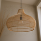 A large woven rattan pendant light in a natural tan color, hanging in a warm, neutral-toned room with a textured beige wall and dried grass decor in the background.