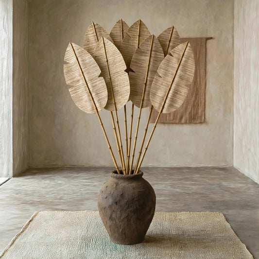A rustic brown ceramic vase filled with large dried palm leaves on bamboo stakes, displayed on a woven rug in a minimalist, earthy-toned room.