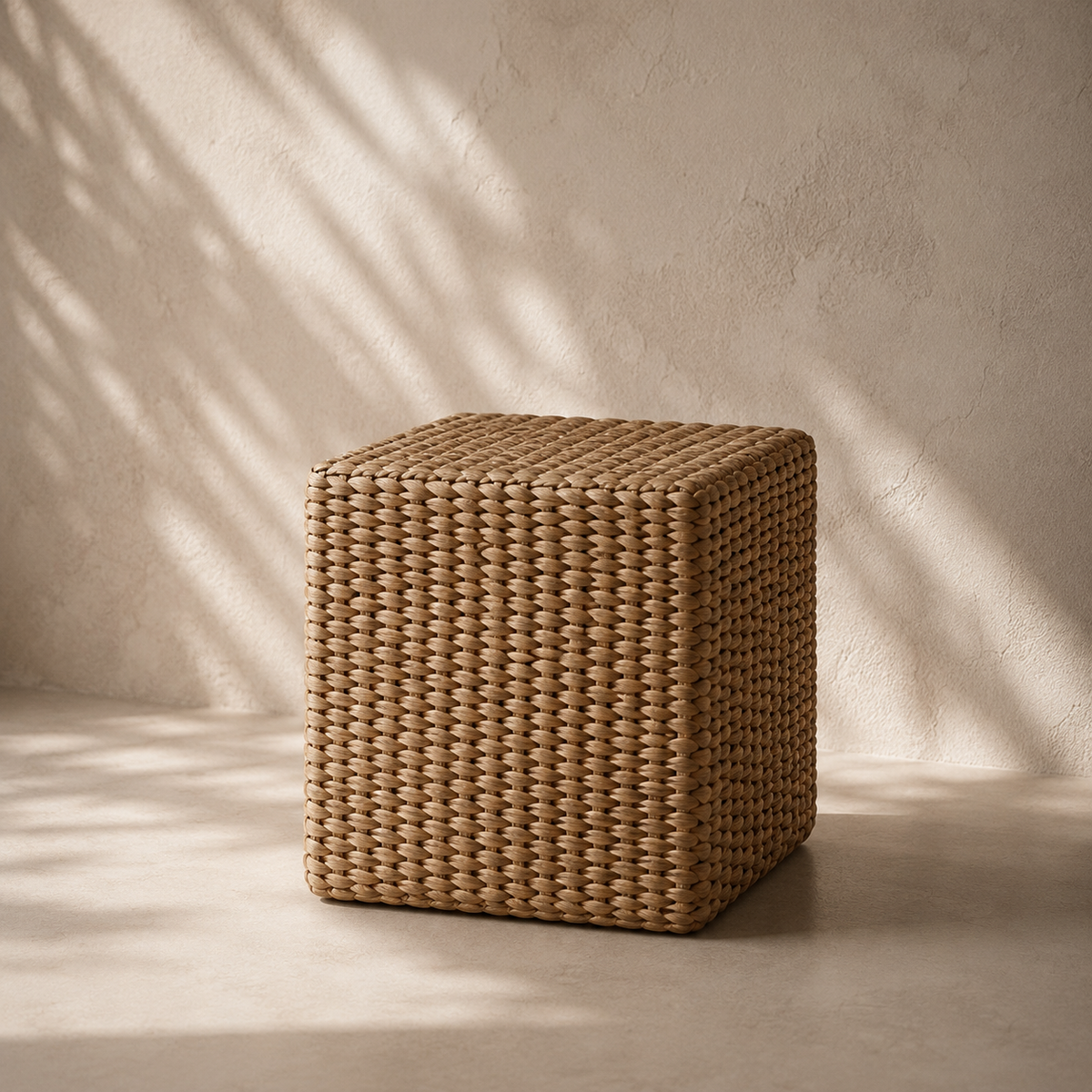 A cube-shaped woven rattan pouf or ottoman in a natural tan color, positioned on a concrete floor against a textured plaster wall with soft leaf shadows cast across it.