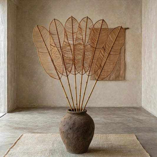 A large rustic brown ceramic vase holding a tall, fan-like arrangement of thin woven reed or straw stems, placed on a natural jute rug in a minimalist concrete room with textured walls.
