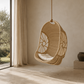 A woven hanging egg chair with a white cushion suspended by a rope in a minimalist, warm-toned interior room with a large window showing an outdoor view.