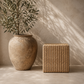 A square woven rattan stool or side table in a natural tan color stands next to a large rustic beige ceramic vase holding an olive branch, set against a textured beige wall with soft leaf shadows.