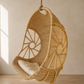 A natural woven rattan hanging egg chair with a cream cushion, suspended by a rope in a minimalist room with warm sunlight and a textured beige wall.