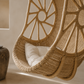 A close-up of a woven rattan hanging egg chair with a white cushion, set against a warm beige wall.