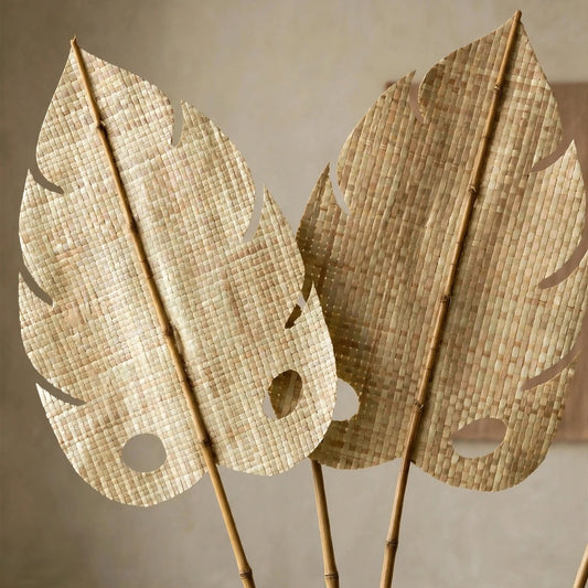 Large decorative woven monstera leaves on bamboo-style stakes, displayed against a neutral textured beige wall.