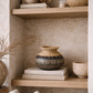 A styled interior shelf scene with natural woven baskets, ceramic vases, stacked books, and dried branches against a textured beige wall.