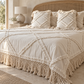 A bohemian-style bed dressed in cream-colored textured bedding, including a fringed quilt, matching pillows, and a woven cane headboard, set in a warm, neutral-toned bedroom.
