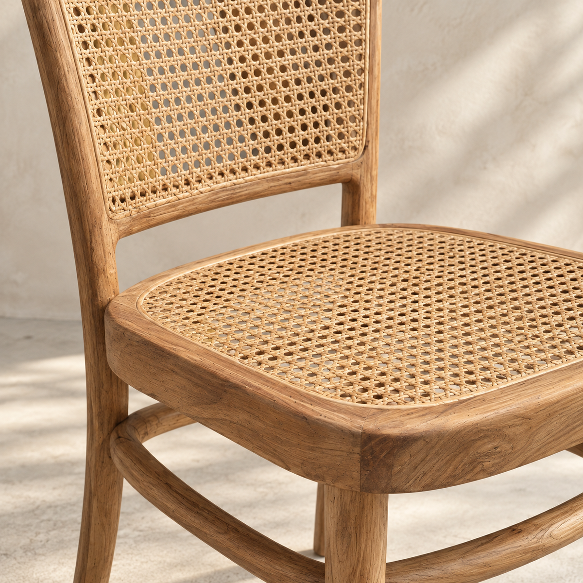 Close-up of a wooden dining chair with a woven cane backrest and seat, set against a textured light beige wall with soft shadow patterns.
