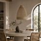 A woven natural fiber pendant light hangs over a round dining table in a warm, sunlit room with wicker chairs, a ceramic vase with greenery, and an arched window.