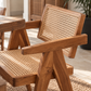 A close-up of a wooden dining chair with a woven cane or rattan seat and back, shown in a warm indoor setting with a table and rug nearby.