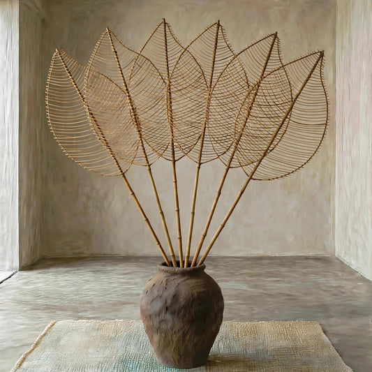 A decorative sculpture resembling a lotus flower, made of many thin bamboo-like stems radiating from a round textured brown vase, displayed on a woven rug inside a minimalist concrete room.
