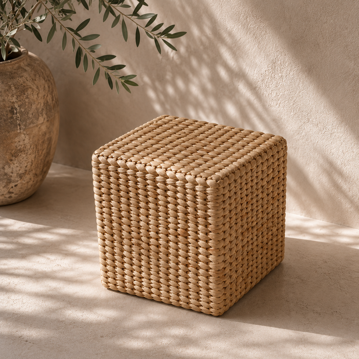 A cube-shaped woven rattan pouf or side table in a natural tan color, styled on a concrete surface with dappled sunlight and shadows, next to a rustic ceramic planter.
