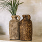 Two brown terracotta vases with a textured surface, displayed against a neutral background with a green plant.