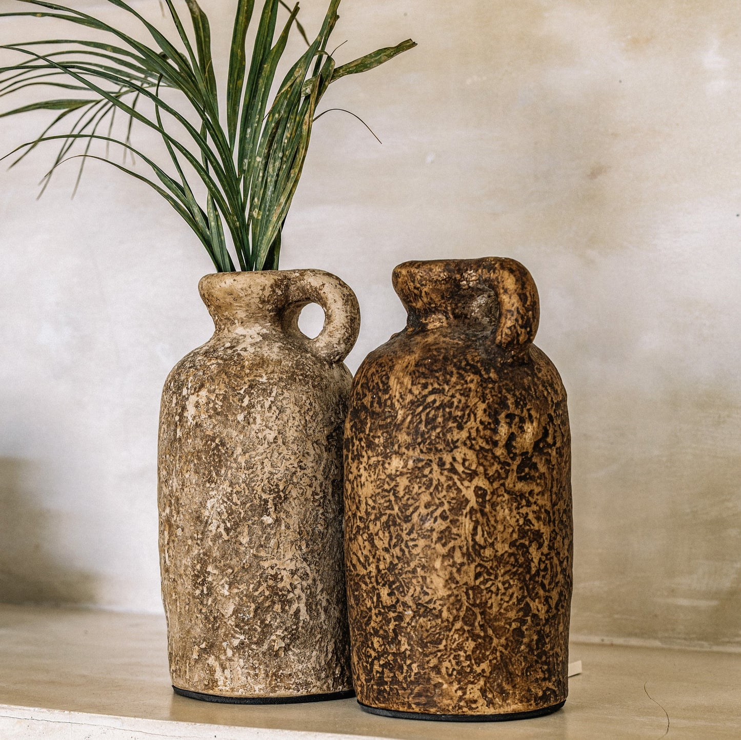 Two brown terracotta vases with a textured surface, displayed against a neutral background with a green plant.