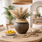 A bohemian-style dining room scene featuring a round wooden table with woven rattan chairs, a large woven pendant light, and a central ceramic vase filled with pampas grass and greenery, styled with lemons and candles.