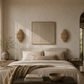 A minimalist bedroom scene with a neatly made bed in neutral beige linens, a textured throw blanket, two bedside tables, wall sconces, and a framed artwork centered on the wall.
